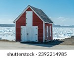 Small photo of An old two story vibrant red barn with double white doors and a hay loft. The roof is made of black cedar shakes. The shed is at the edge of an icy shore. The harbor is filled with thick slob ice.