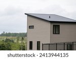 The exterior corner of a modern wooden tiny house. It has grey vinyl walls with black trim, closed glass windows, slanted flat roof, black metal backyard fence and lush green views of the countryside.