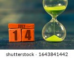 Small photo of july 14th. Day 14 of month,Handmade wood cube with date month and day and hourglass with green sand. Time passing away. artistic coloring. summer month, day of the year concept