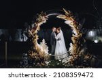 A stylish groom and a beautiful bride in a long dress are standing at night near a reed arch decorated with lamps and garlands. Wedding photography, portrait.