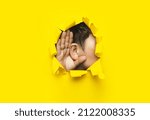 Small photo of Close-up of a man's ear and hand through a torn hole in the paper. Yellow background, copy space. The concept of eavesdropping, espionage, gossip and tabloids.