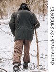 Small photo of An old, elderly forester, a homeless beggar man in dirty torn clothes walks through the forest with a wooden stick in search of food, shelter in the cold snowy winter. Photography, poverty concept.
