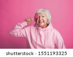 Positive, happy and cheerful old woman posing on camera alone. Stand and cover eye with lollipop. Wear stylish hoody. Isolated over pink background
