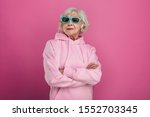 Cool old woman in funny green sunglasses posing on camera. Hands crossed. Wear pink hoody. Stylish modern grandma. Isolated over pink background