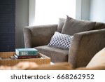 Low angle cushioned gray fabric upholstered living room seat with blue and white patterned throw pillow below highlighted window with stack of coffee table books atop tray on ottoman foot rest 