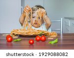 Small photo of Funny baby child getting messy eating spaghetti with tomato sauce from a large plate, by itself with his hands, at home