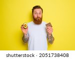 Small photo of Doubter man with beard and tattoos holds avocado and chocolate