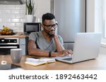 Small photo of Tired african american male employee sit at the table in home office, using laptop, smiling. Focused exhausted businessman work on own project, having a tense and ache in back, needing a rest