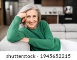 Small photo of Portrait of charming senior woman with grey hair, smiling mature female sits on the couch at ease and looks at the camera. Beautiful carefree middle-aged woman at home