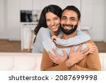 Happy lovely young Indian couple together at home, ethnic young wife hugging from behind her husband, sitting and resting on sofa in modern apartment, portrait of romantic multiracial couple in love