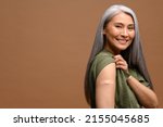 Small photo of Smiling senior grey-haired woman with a medical patch after vaccination isolated on brown background, protecting hand with bandage after injection. Healthcare and medicine concept