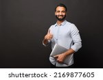 Small photo of Indian male entrepreneur in casual shirt carrying laptop and showing thumb up isolated on black, bearded businessman with laptop looking at the camera and smiles, ok gesture