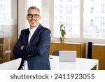 Optimistic gray-haired old hand mature businessman in formal wear standing indoors, smiling senior male entrepreneur 50s looks at the camera leaned at the office desk, male employee with crossed arms