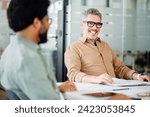 Small photo of An affable mature man with glasses engages in a friendly discussion with a coworker, showcasing the warm and approachable nature of a collaborative office meeting
