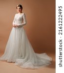 Small photo of Luxury wedding dress. Fashionable bridal look. Ball gown with tender french lace and beads, long sleeves, lush tulle skirt. Beautiful young blonde bride in studio on brown background. Marriage concept