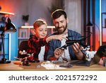 Caucasian man and little boy repairing modern quadcopter while sitting at table. Happy satisfied father teaching son different man's work at home. Parenting concept.