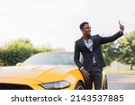 Positive african man in suit having video call on smartphone while standing near luxury sport car outdoors. Businessman standing near yellow auto, smiling and waving during online conversation