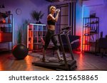 Small photo of Beautiful woman drinking water from bottle, exercising, having workout on a treadmill in living room. Happy female running, having cardio training at evening time at home, healthy lifestyle concept.