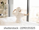 Small photo of Back view of young lady in bath towel and white robe pulling hands aside while sitting in front of panoramic window in airy bedroom. Cheerful woman relieving tension before starting new day at home.