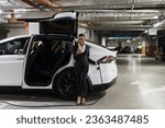 Small photo of Full length view of elegant woman dialing number on smartphone while leaning against electric vehicle indoors. African office worker getting unlimited supply of renewable energy in parking lot.