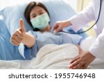 Happy Asian female patient wears a mask, lies on the bed, and raises her thumb up. When a doctor uses a stethoscope to listen to the lungs. Concept of believe in treatment And insurance coverage