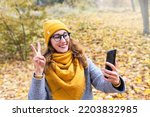 Happy smiling young woman in a yellow hat and yellow scarf shows victory sign talking on video call in the autumn park. Warm autumn day. Autumn mood.