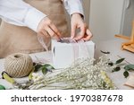 A florist decorates a gift box with flowers and a ribbon on a white desktop. Only the hands are in the frame