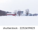 Winter landscape featuring a patrimonial white clapboard farm house with pitched shingled roof, red barn and other outbuildings in land covered in fresh snow, Saint-Vallier, Quebec, Canada