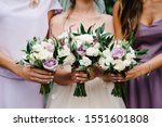 The bride and bridesmaids in an elegant violet dress is standing and holding hand bouquets of pastel pink flowers and greens at nature. Young beautiful girls holds a wedding bouquet outdoors.
