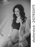 Small photo of Belly of pregnant woman. Female waiting for newborn baby on white background. Young pregnant girl holding belly and caring about health. Pregnancy motherhood procreation concept. Black and white photo