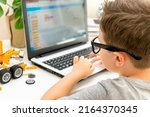 Smart little boy student in glasses builds and programs a robotic vehicle codes an electronic toy. Child using laptop in science class, his hands typing on keyboard. Programming lesson. Rear view.