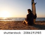 Back view young adult slim sporty female surfer girl with surfboard sitting on sand at ocean coast wave against warm sunrise or sunset sun. Sport healthy carefree freedom lifestyle vacation concept