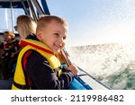 Portrait of cite little blond happy excited smiling caucasian boy wear lifevest enjoy sailing on motor boat sea against blue sky and water splash wave sun backlit. Summer travel vacation recreation