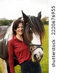 Small photo of A woman smiles for a portrait beside her skewbald Irish Gypsy Cob horse in a grassy field