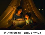 Asian family mom and girl happy at night time in bedroom at home before bed time reading fantasy bedtime story book together in kid tent with dim yellow light