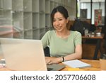 Photo of beautiful Asian woman working with a computer laptop at the wooden table over the cozy living room as a background.