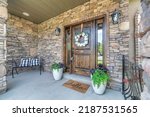 Small photo of Entrance of a house with stone walls lockbox on the wooden front door with two sidelights in Utah. Porch of a house with white wreath and two potted plants on the door and a bench on the left.