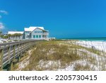 Boardwalk and beach house near the white sand shore of a beach at Destin, Florida. There are grasses on the white sand and a view of a gray house with terrace at the back.