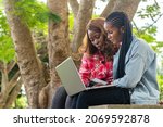 two female african students sitting on a park bench using a laptop to check something online