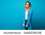 Beautiful young woman in round glasses, beige blouse and trendy blue coat isolated at blue background. Model girl in blouse, blue coat and modish eyewear over blue background. Copy space. Spring look