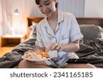 Asian teenager girl picking up snacks from plate,eating junk food,potato chips and popcorn on her bed at night,enjoy watching movie on TV,eat late and stay up late,health care and nutrition concept