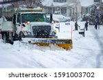 Front view of city services snow plow truck with yellow push blade clearing covered roads after heavy winter snow fall and passing by children bundled in warm clothes playing while flakes still fall.