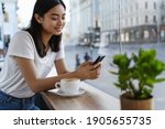 Summer girl sitting in cafe with cup of coffee, messaging on smartphone. Young woman using mobile phone in restaurant while drinking cappuccino near window.