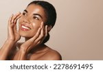 Small photo of Young african american girl with clear, glowing facial skin, washes her face with cosmetic product, smiling and looking up, enjoys smooth feeling after hydrating serum, face mask