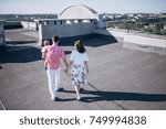 Small photo of Woman and man walking on the rooftop. They are holding hands each other. Man holds his little son on the hand. They enjoy morning sun and amazing view. They are wearing tricksy light summer clothes.