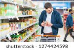 Small photo of At the Supermarket: Handsome Man Uses Smartphone and Looks at Nutritional Value of the Canned Goods. He's Standing with Shopping Cart in Canned Goods Section.