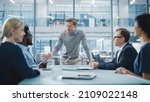 Small photo of Modern Office Meeting Room: Handsome Executive Director Stands in the Head of the Conference Table Leans on it and Delivers Eloquent Speech to a Group of Businesspeople and Investors.