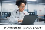 Small photo of Hospital Doctors Office: Portrait of Young Promising Black Physician Works on Laptop Computer. Female Medical Professional in White Lab Coat Looking for Patient Treatment While Sitting at Her Desk