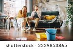 Small photo of Roof is Leaking or Pipe Rupture at Home: Panicing Couple In Despair Sitting on a Sofa Watching How Water Drips into Buckets in their Living Room. Catastrophe, Distaster and Financial Ruin