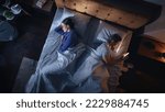 Small photo of Top View Bedroom Apartment: Man Uses Smartphone in Bed at Night When His Female Partner Trying to Fall Asleep. Couple After Fight, Argument. Addictive World of Social Media, Doom Scrolling, Fake News.